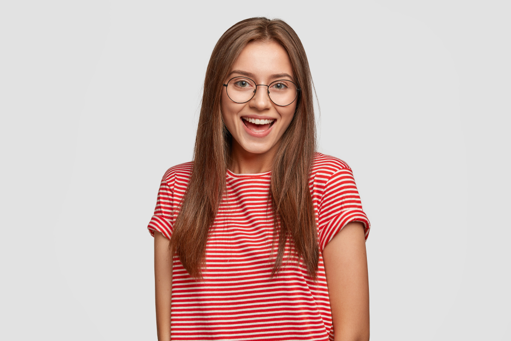Joyful Dark Haired Woman With Toothy Pleasant Smile, Wears Round Transparent Glasses, Striped T Shirt, Being In Good Mood, Recieves Positive News From Interlocutor, Isolated Over White Background